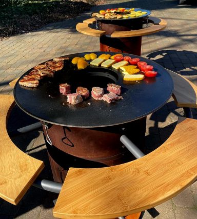 Grill mit Fleisch und Gemüse auf einer runden Edelstahlplatte, Sitzgelegenheiten drumherum.